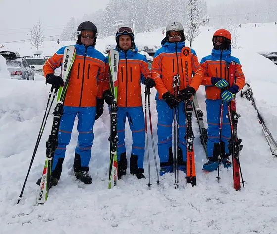 Vier Skifahrer in orange-blauen Teamjacken mit Rennski